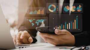 Pessoa utilizando notebook e smartphone simultaneamente, com diversos gráficos de barras, pizzas e mapas holográficos flutuando sobre a mesa, representando a análise de dados processados.