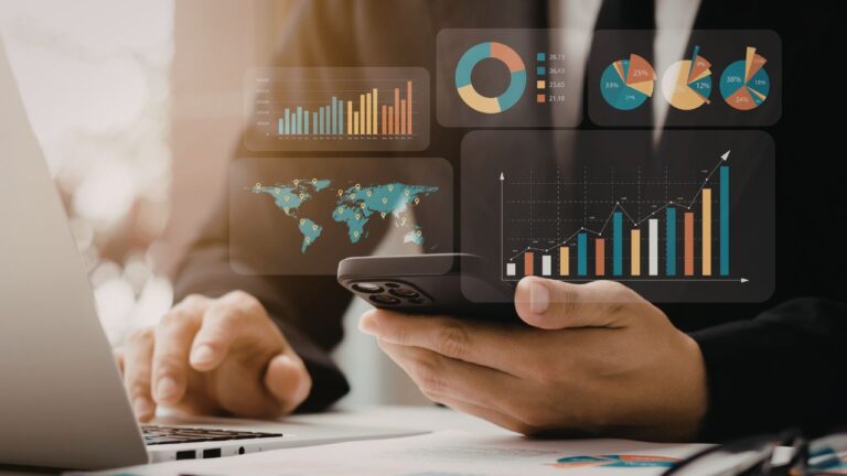Pessoa utilizando notebook e smartphone simultaneamente, com diversos gráficos de barras, pizzas e mapas holográficos flutuando sobre a mesa, representando a análise de dados processados.