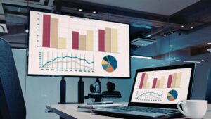 Escritório moderno com um laptop em primeiro plano e uma grande tela de TV ao fundo, ambos exibindo o mesmo dashboard com gráficos de barras coloridas, gráficos de linha e um gráfico de pizza.