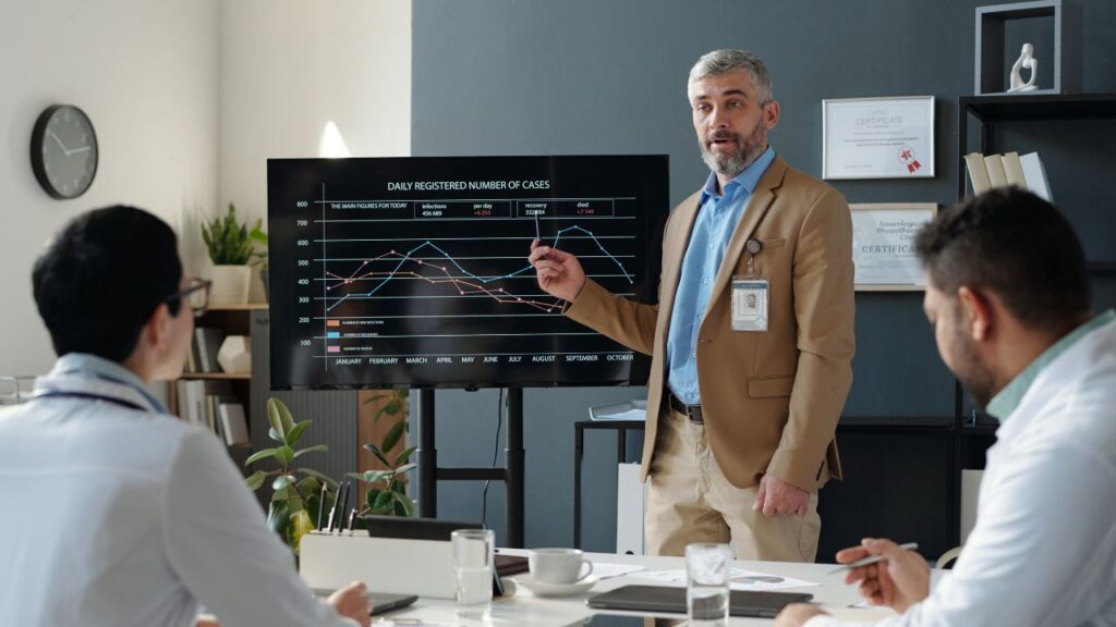 Homem de barba com terno cáqui em pé ao lado de uma tela de TV, apontando para um gráfico de linhas que mostra o número diário de casos registrados, enquanto explica os dados para uma audiência em uma sala de reunião.