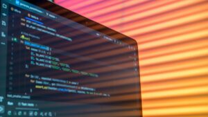 Close-up em ângulo lateral de um laptop exibindo um editor de código (IDE) em fundo escuro com linhas de código de programação coloridas (como JavaScript/Python), funções e chaves. A luz ambiente é quente, em tons de azul e laranja, com reflexos suaves na tela. Ao fundo, uma textura de persianas listradas laranja e amarela.