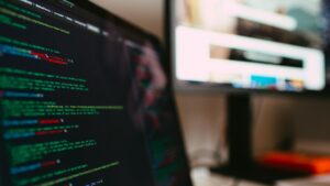 Close-up em ângulo lateral de um notebook à esquerda exibindo um editor de código em modo escuro com linhas de código de programação coloridas (como HTML, CSS ou JavaScript). Ao fundo e à direita, um monitor de desktop maior exibe uma interface gráfica de site desfocada com elementos de layout visíveis sob luz ambiente suave.