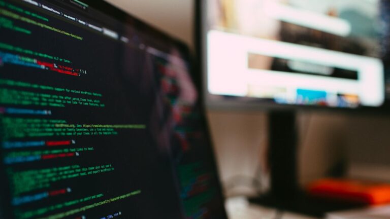 Close-up em ângulo lateral de um notebook à esquerda exibindo um editor de código em modo escuro com linhas de código de programação coloridas (como HTML, CSS ou JavaScript). Ao fundo e à direita, um monitor de desktop maior exibe uma interface gráfica de site desfocada com elementos de layout visíveis sob luz ambiente suave.