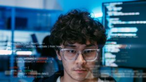 Foto em primeiro plano de um jovem de óculos transparentes e cabelos cacheados, com expressão focada. Sobrepostas à cena, há linhas translúcidas de código de programação JavaScript (com destaque para um componente React e chamadas axios), ícones binários (01001) e uma interface de dashboard. Ao fundo, um monitor exibe mais código sob luz azul.